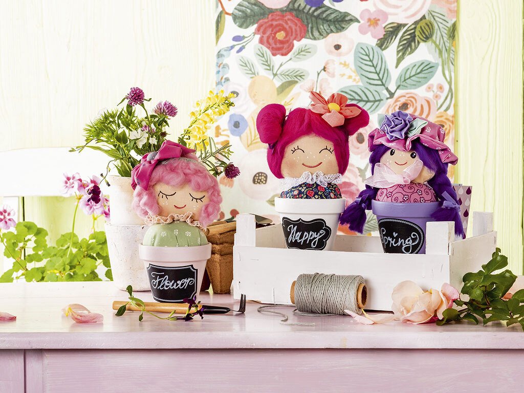 Drei Nadelkissen als Stoffpuppen in bemalten Blumentöpfen auf einem Tisch mit Blumen und Nähgarn