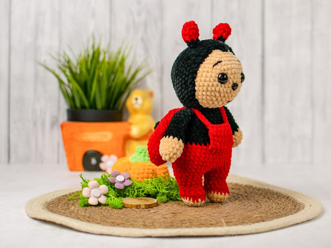 Gehäkelte Amigurumi-Marienkäfer-Puppe mit roten Latzhosen auf runder Matte vor Topfpflanze