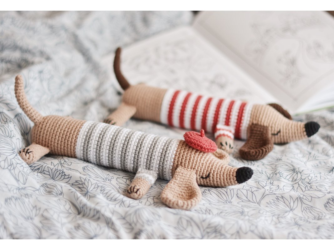Zwei gehäkelte Dackel-Amigurumi in gestreiften Pullovern, einer mit roter Baskenmütze, auf geblümter Decke.