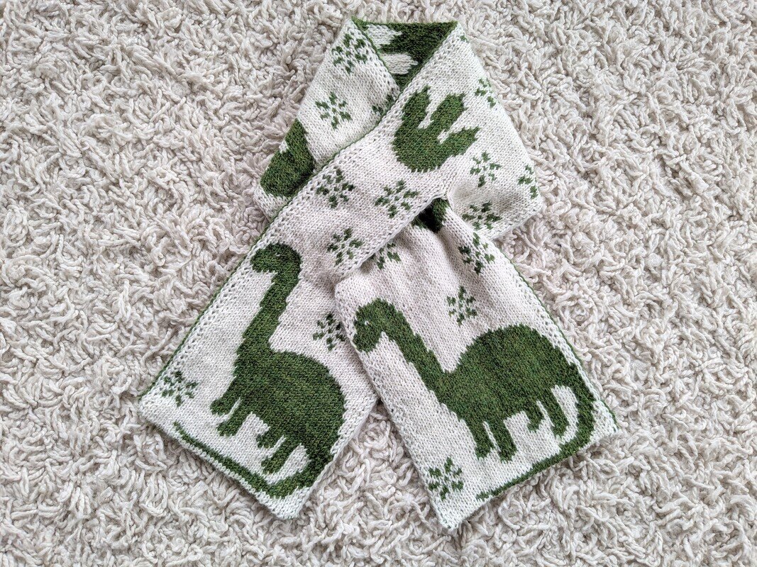 Child-sized knitted scarf with green dinosaur motifs and small star repeats on a cream background