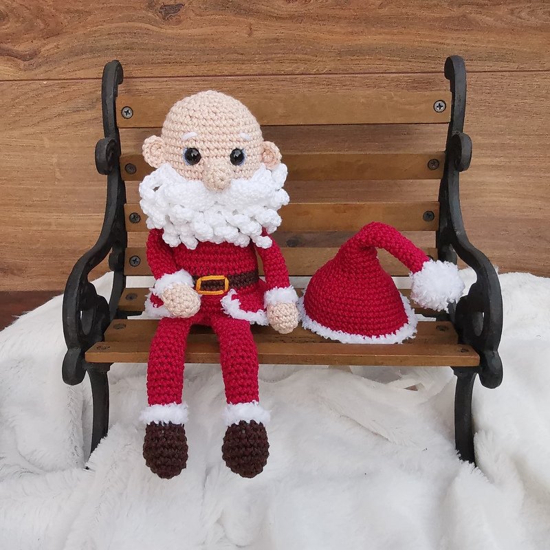 Gehäkelte Amigurumi-Weihnachtsmann-Puppe mit weißem Bart und rotem Anzug auf einer Holzbank, die rote Zipfelmütze liegt daneben