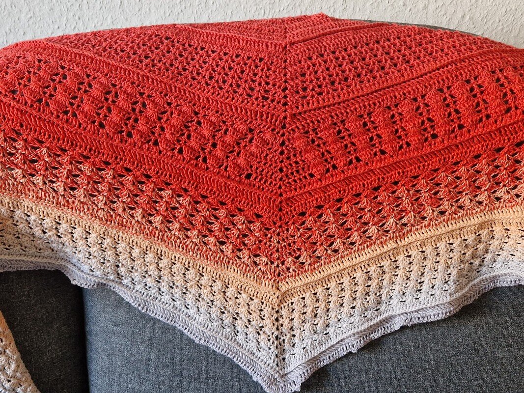 Gehäkeltes Dreieckstuch in Koralle, Beige und Grau mit Ombré-Farbverlauf und Lochmuster, auf einem grauen Sofa drapiert.