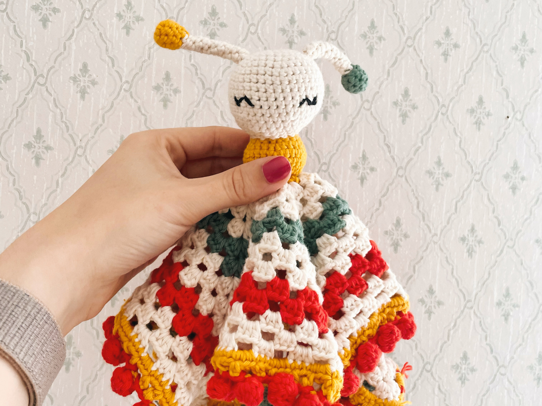 Detailansicht eines gehäkelten Schmusetuchs im Käfer-Design mit buntem Granny-Square-Muster, von einer Hand präsentiert