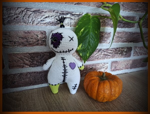 Gehäkelte weiße Amigurumi-Puppe mit gestickten Nähten neben kleinem Kürbis