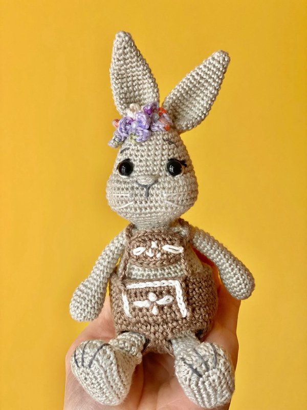 Gehäkelter Amigurumi-Hase mit brauner Latzhose und Blümchen am Ohr, gehalten in einer Hand vor gelbem Hintergrund.