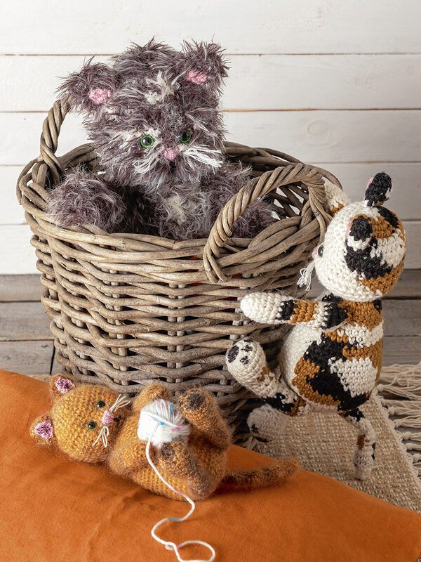 Drei gehäkelte Katzen-Amigurumi: graue Katze im Weidenkorb, orange Katze auf Kissen mit Wollknäuel, gefleckte Katze daneben.