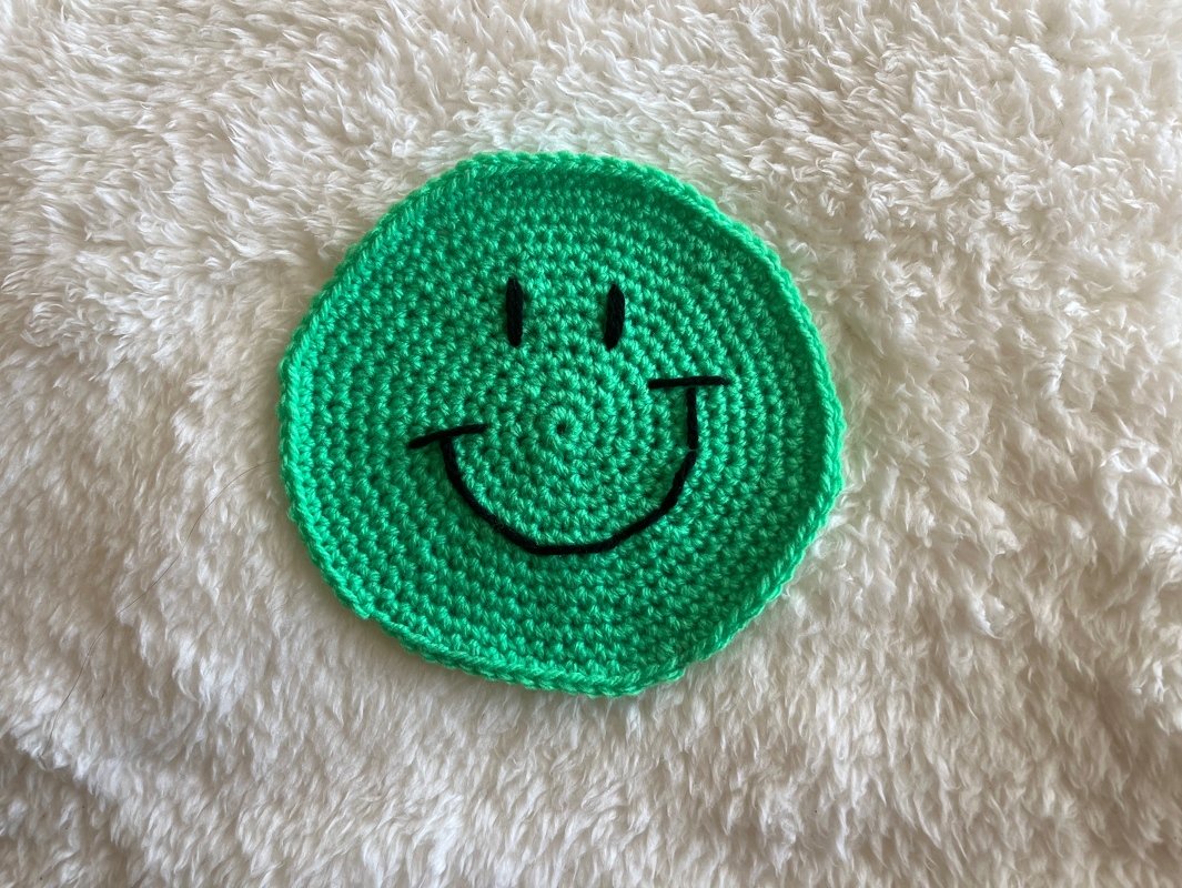 Runder grüner, gehäkelter Smiley-Untersetzer auf weißem flauschigem Stoff.