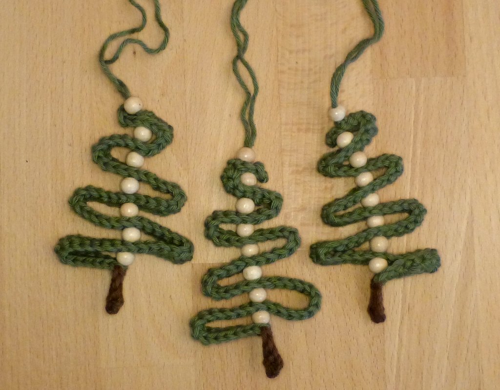 Drei gehäkelte grüne Tannenbaum‑Anhänger mit weißen Perlen und braunen Stämmen auf Holz
