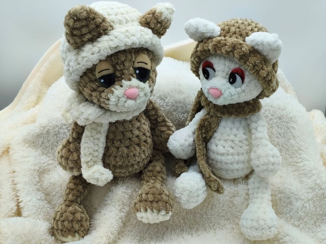 Zwei gehäkelte Amigurumi-Kätzchen in Braun und Creme mit Mützen und Schals auf flauschiger Decke.