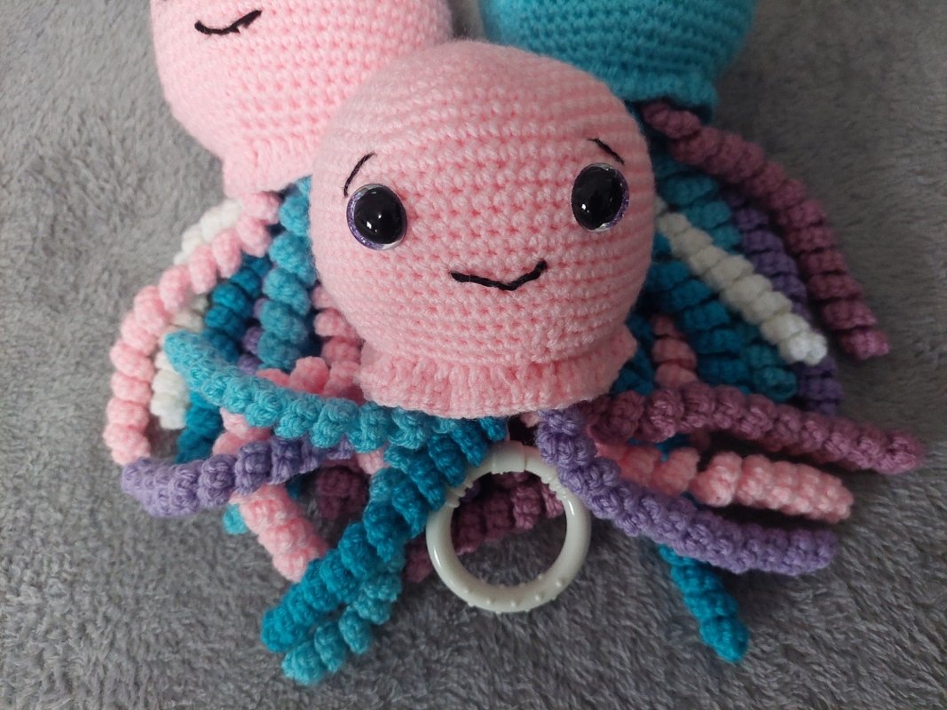 Gehäkelter rosa Oktopus mit bunten spiralförmigen Tentakeln und weißem Beißring auf grauem Untergrund.