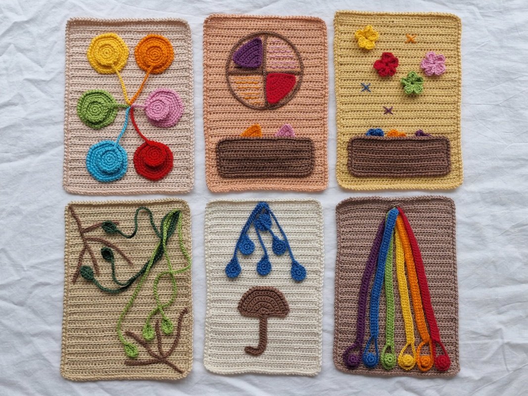 Sechs farbige, handgehäkelte rechteckige Tafeln mit verschiedenen kindgerechten Motiven auf weißem Stoff angeordnet.