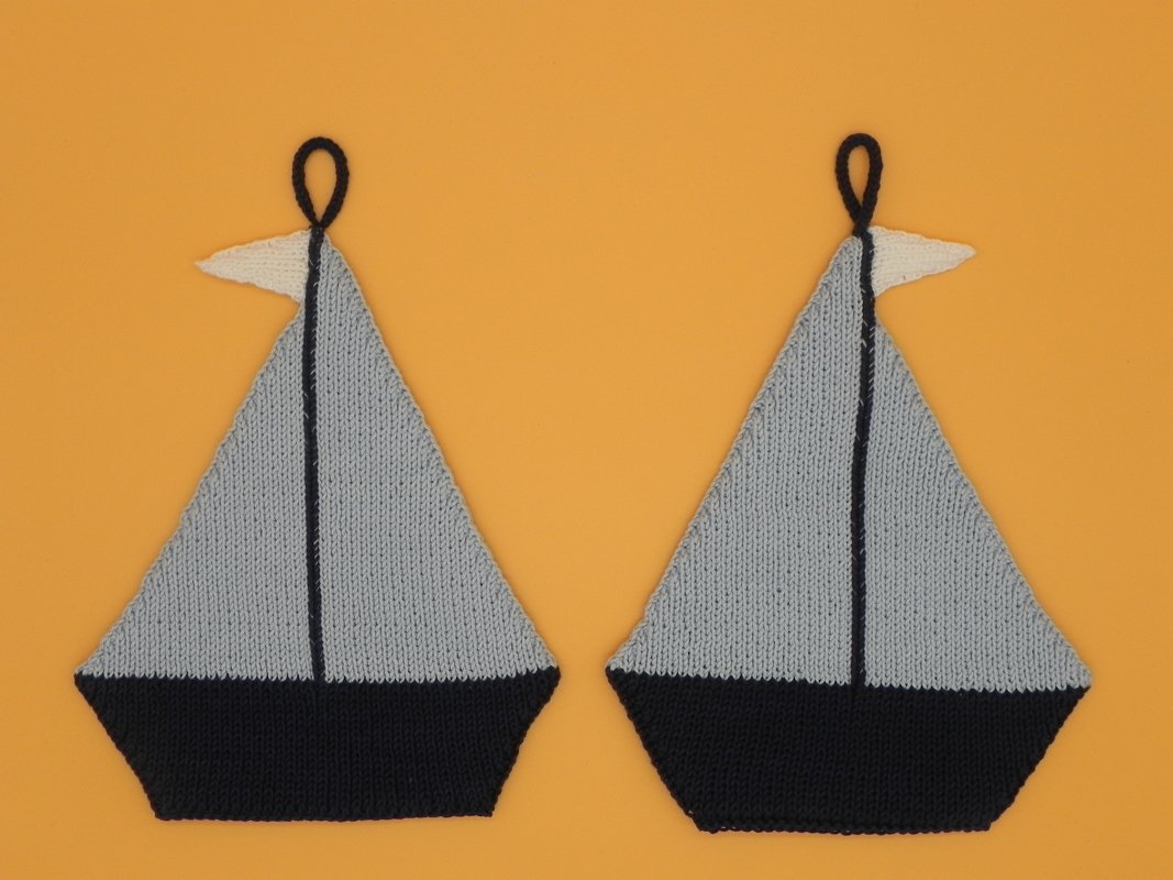 Zwei gestrickte Segelboot-Topflappen mit hellgrauen Segeln, dunklen Rümpfen, weißen Wimpeln und Aufhängeschlaufen auf orangefarbenem Hintergrund.