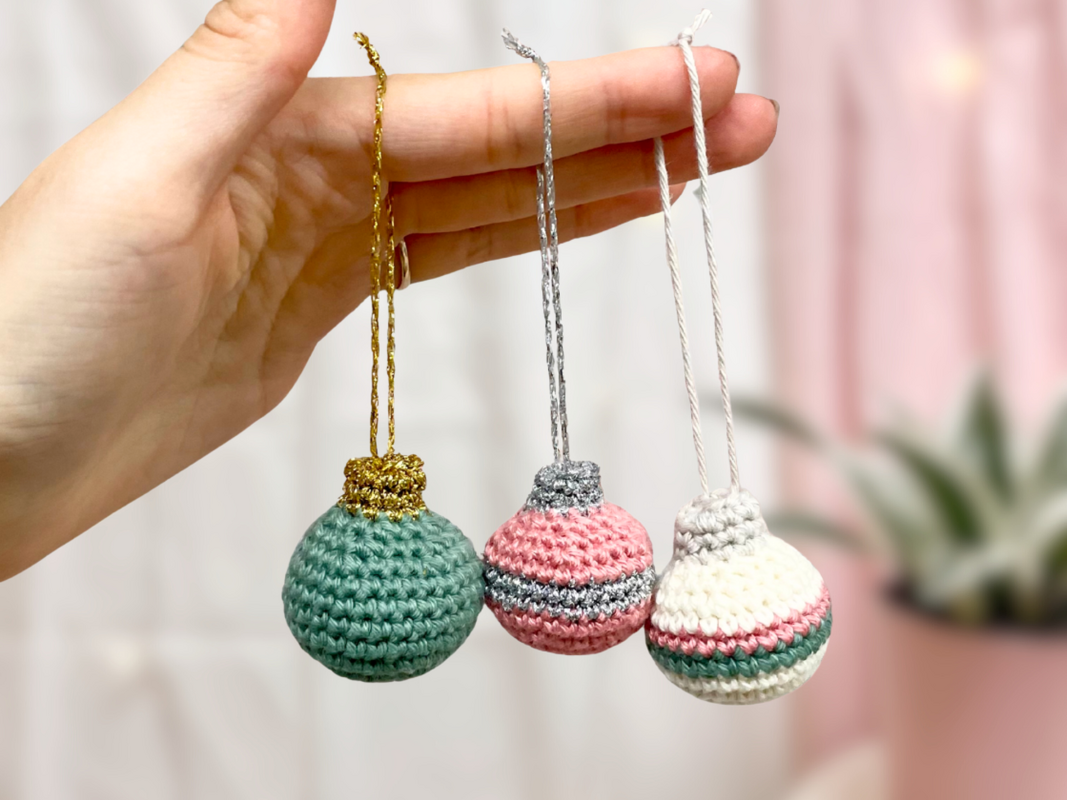 Hand hält drei gehäkelte Weihnachtskugeln in Pastell mit goldener, silberner und weißer Aufhängeschlaufe