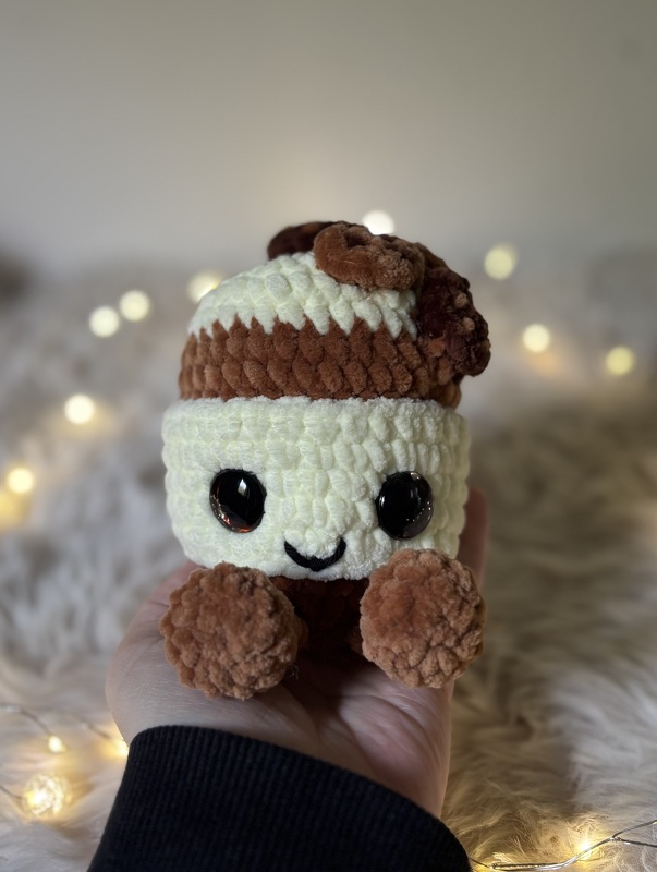 Hand hält ein gehäkeltes Amigurumi in Cremeweiß und Braun mit großen Augen und lächelndem Mund.