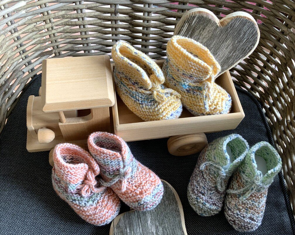 Drei Paare handgestrickter Babyschuhe in Pastellgelb, Pfirsich und Hellgrün mit Bindebändchen, arrangiert im Weidenkorb neben Holzspielzeug und Holzherzen.