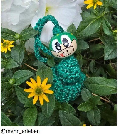 Gehäkelter grüner Frosch-Anhänger mit bemalter Holzkugel als Kopf zwischen Pflanzen und gelben Blüten