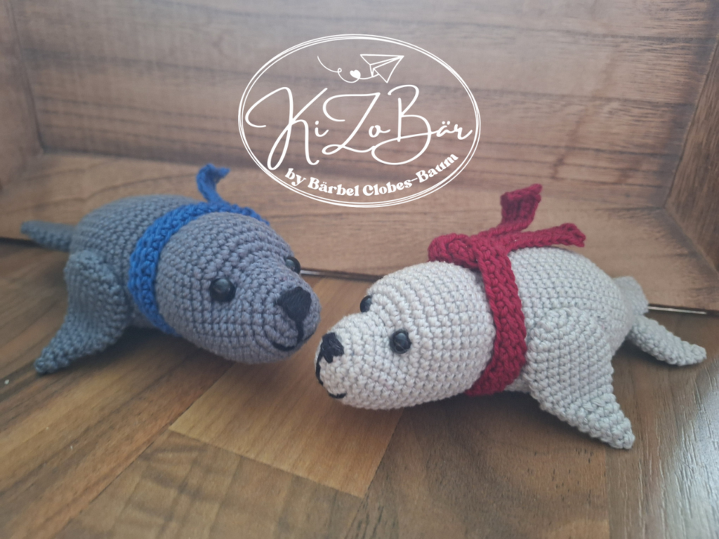 Zwei gehäkelte Seehunde aus grauem Garn mit rotem und blauem Halsband auf Holzuntergrund