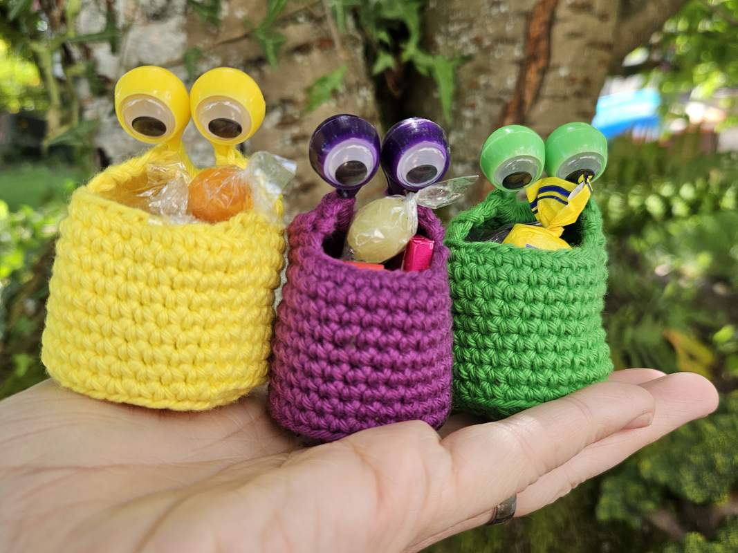 Drei gelbe, lila und grüne gehäkelte Mini-Beutel mit Kulleraugen auf einer Hand.