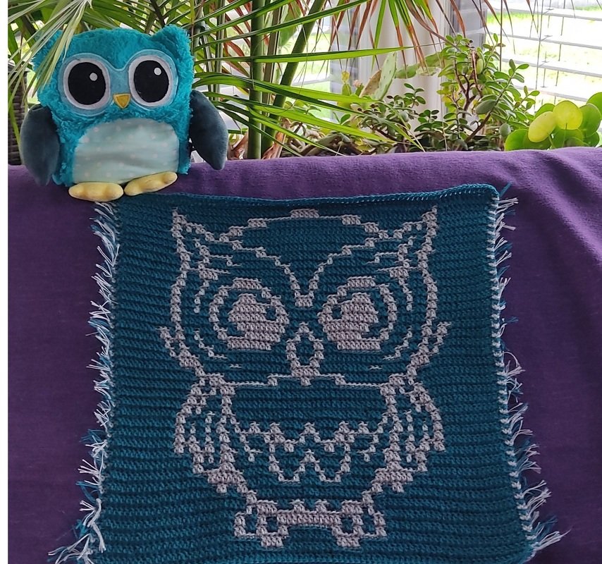 Gehäkeltes Quadrat in Türkis mit grauer Pixeleule auf violettem Stoff, neben einer Plüscheule und Zimmerpflanzen.