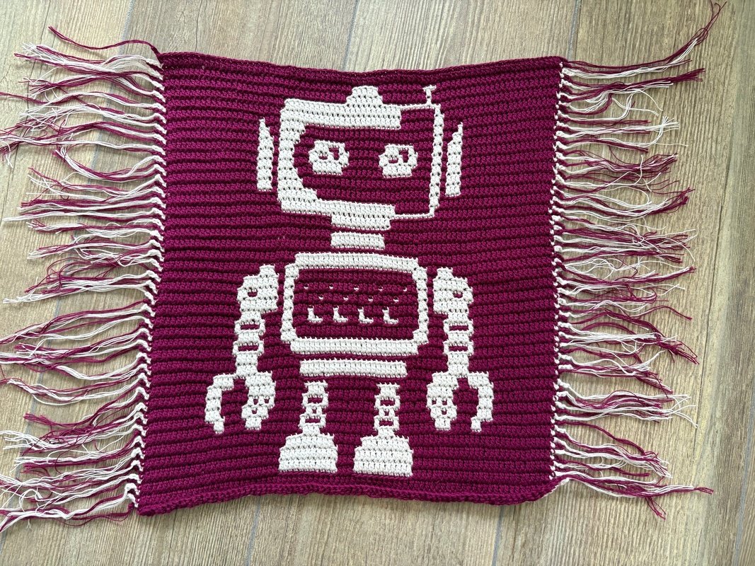 Handgehäkeltes Rechteck mit weißem Pixel‑Roboter auf magentafarbenem Hintergrund und Fransen