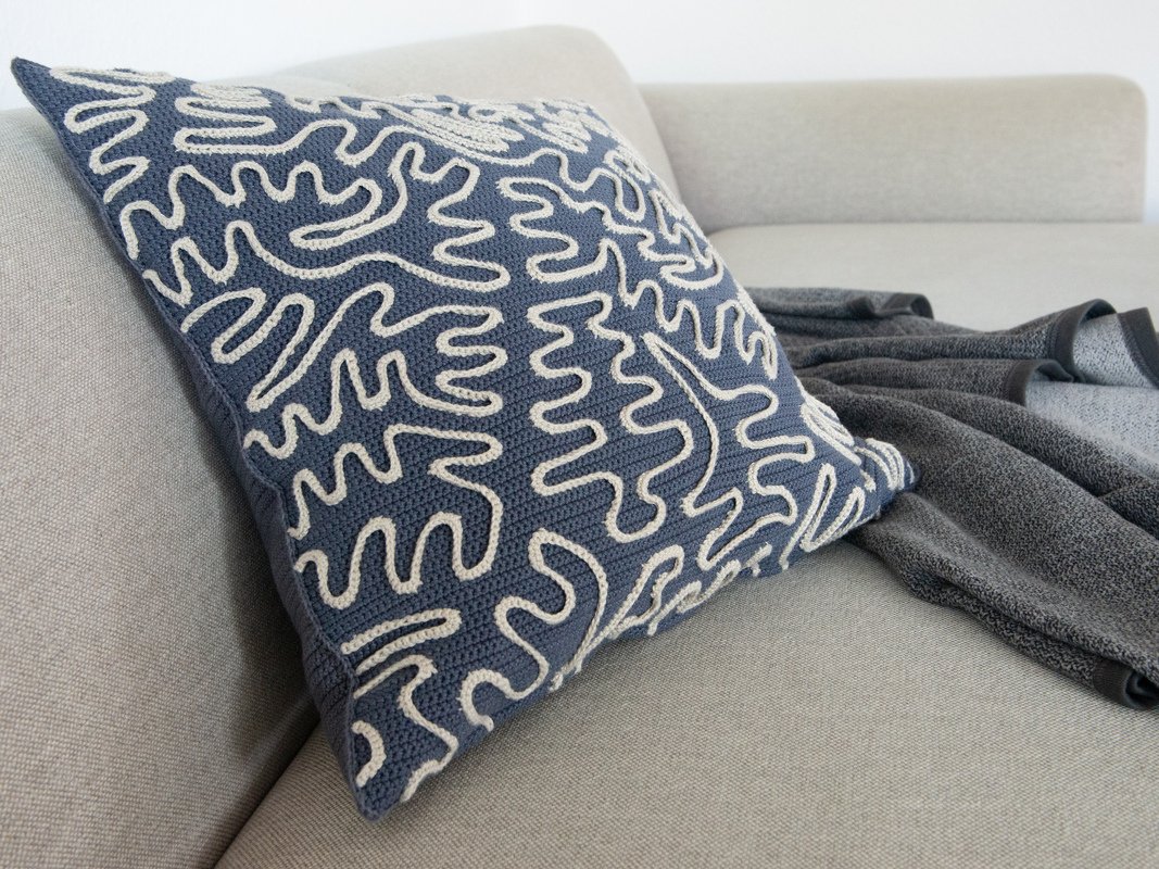 Square blue crocheted pillow with off-white abstract squiggle pattern on a light beige sofa.