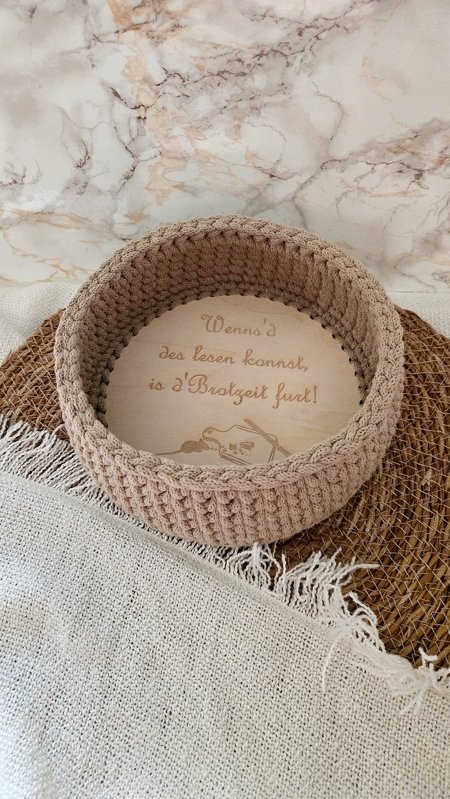 Beige gehäkelter Korb mit hellem Holzboden, darauf eine bairische Gravur und ein Froschmotiv. Der Korb steht auf einer geflochtenen Matte und einem hellen Tuch.