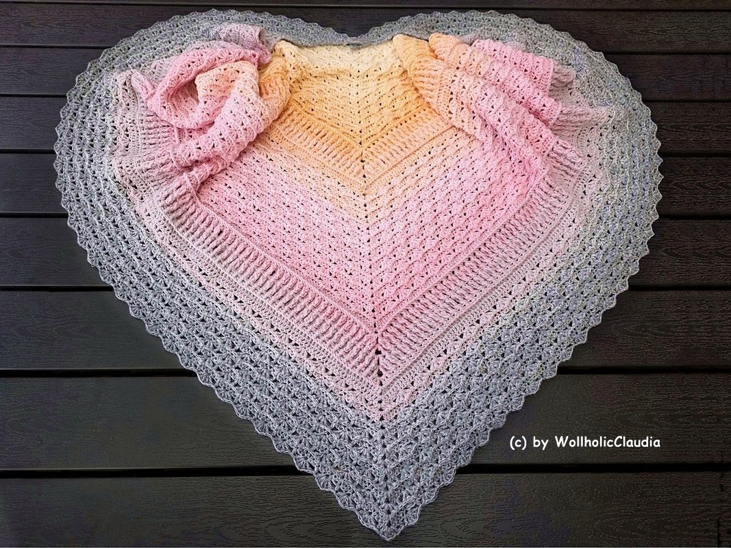 Herzförmiges Häkeltuch mit Pastellverlauf (Gelb, Rosa, Grau) auf dunkler Holzfläche