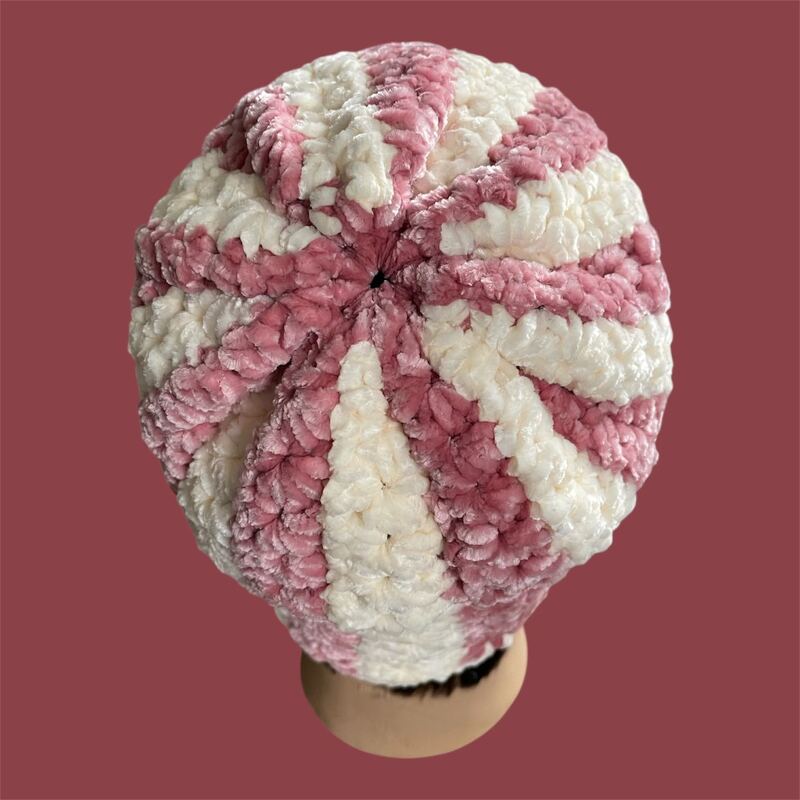 Draufsicht auf eine gehäkelte Beanie-Mütze mit rosa und cremefarbenen, radiär angeordneten Streifen und plüschiger Textur.