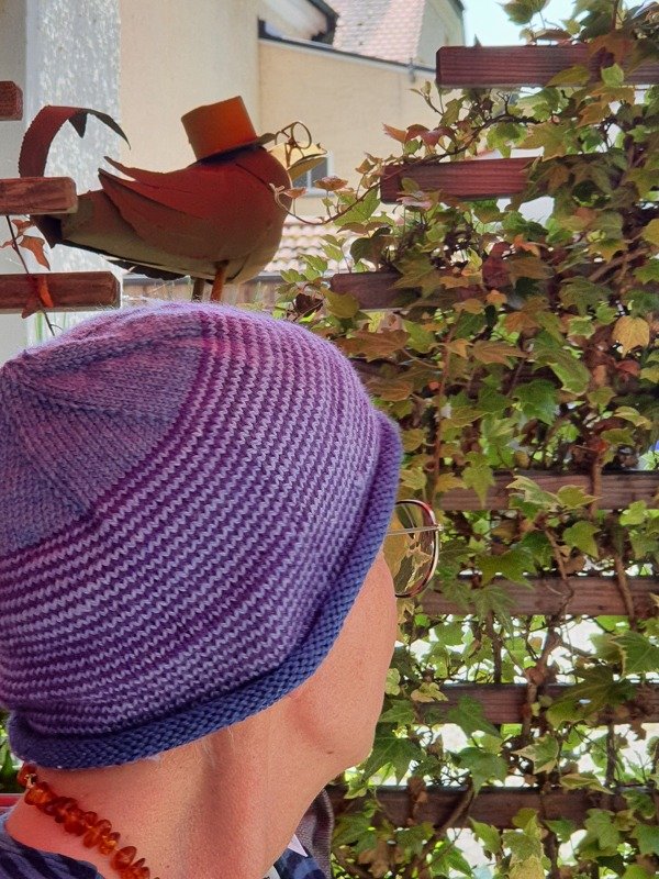 Rückansicht einer Person mit gestrickter Mütze in Lila- und Blautönen vor Efeu und Metallvogel-Dekoration.