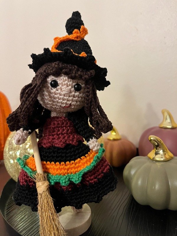 Gehäkelte Amigurumi-Hexe mit schwarzem spitzem Hut, braunen Haaren, mehrfarbigem Kleid und kleinem Reisigbesen, vor dekorativen Kürbissen
