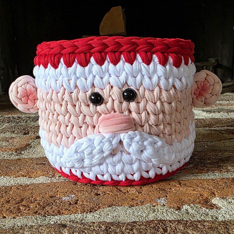 Runder, gehäkelter Korb mit Weihnachtsmann-Gesicht, roter Krempe und weißem Bart auf Ziegelboden.