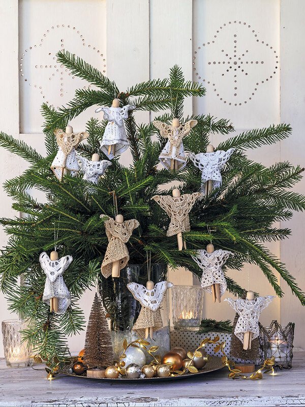 Gehäkelte Engel in Weiß und Beige hängen an grünen Tannenzweigen, umgeben von Kerzen und goldenen Kugeln