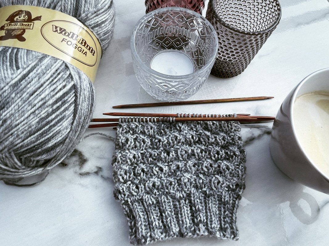 Nahaufnahme eines grauen Strickstücks mit Nadeln, Garnknäuel, Gläsern und Kaffeetasse auf Marmor