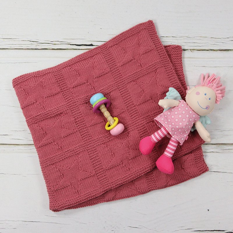 Altrosa gestrickte Babydecke mit quadratischem Texturmuster, daneben Rassel und Stoffpuppe auf hellem Holz.