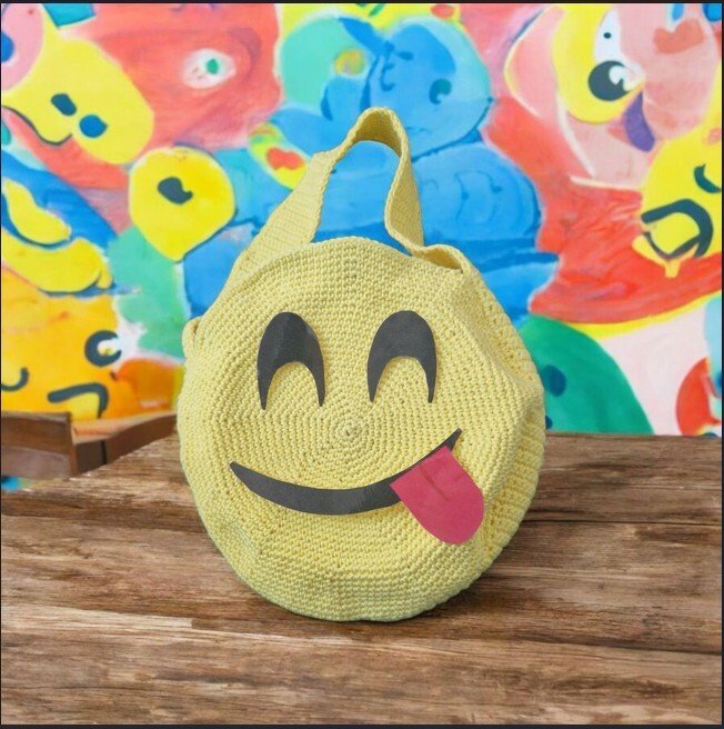 Runde, gelbe Häkeltasche mit lachendem Emoji-Gesicht und herausgestreckter Zunge auf Holztisch