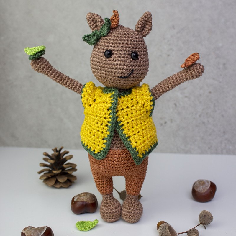 Braune gehäkelte Amigurumi-Tierfigur mit gelber Weste und grüner Umrandung, hält kleine Blätter; daneben Kastanien, Eichel und Tannenzapfen.