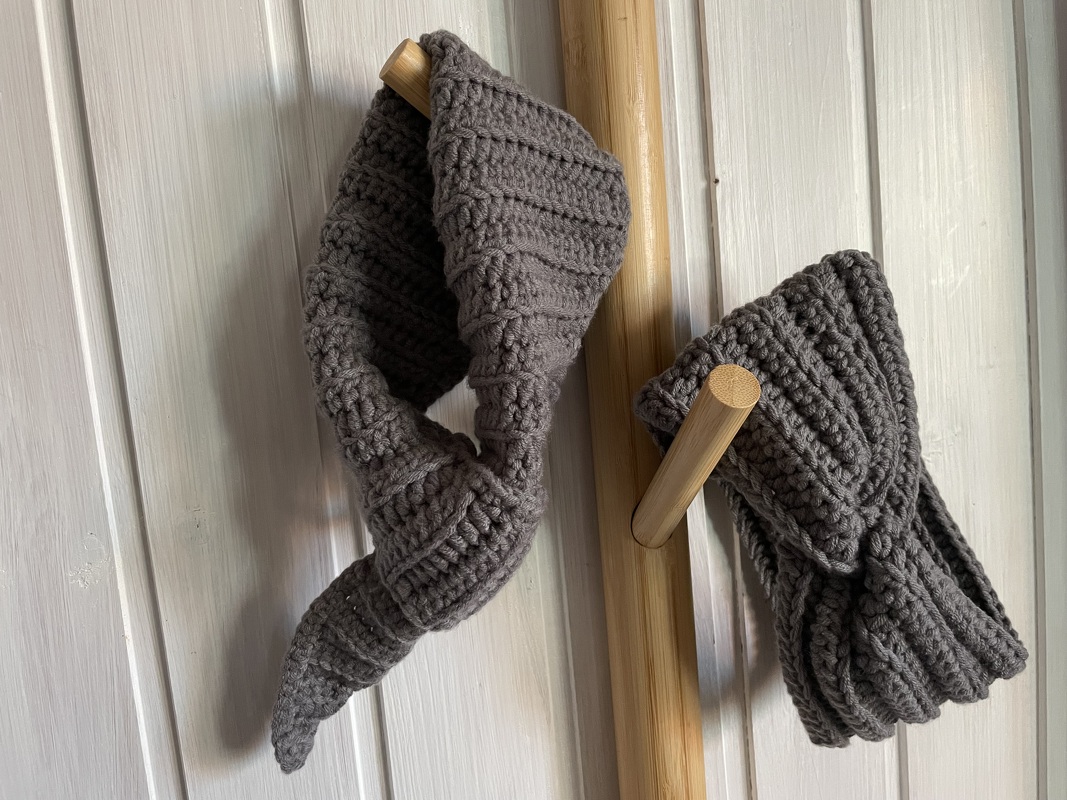 Zwei gehäkelte graubraune Stirnbänder für Kinder an Holzhaken vor weißer Holzwand