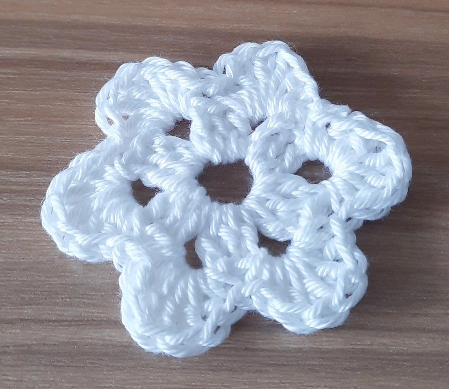 Kleine weiße Häkelblüte mit sechs runden Blütenblättern auf hellem Holz