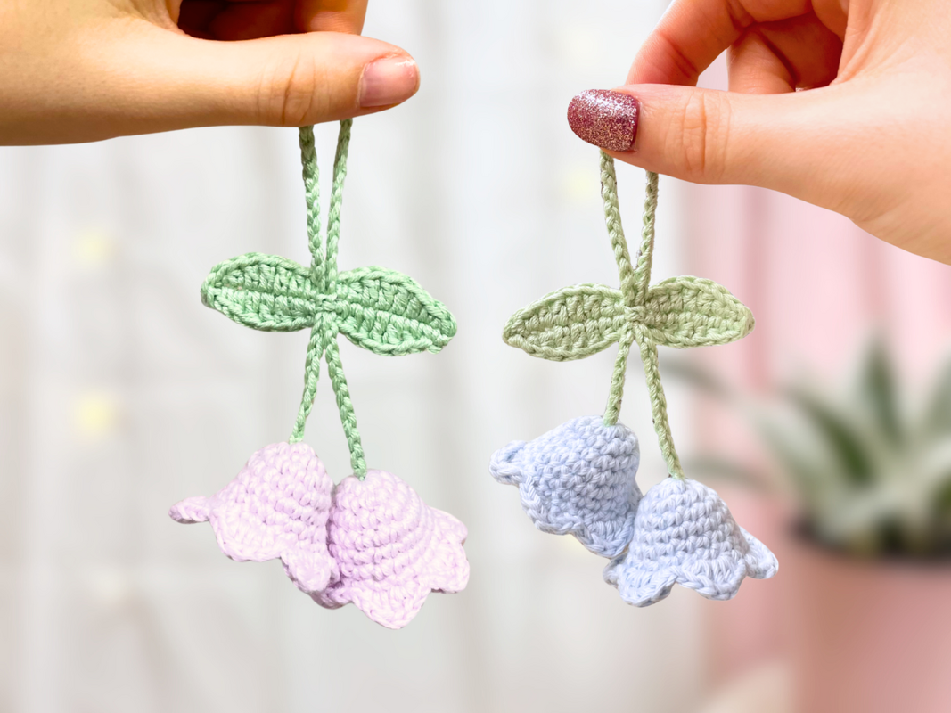 Zwei gehäkelte Glockenblumen‑Schlüsselanhänger in Pastell mit grünen Blättern, von Händen gehalten