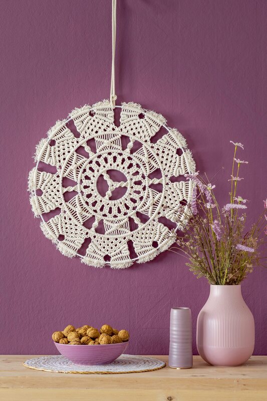 Runder cremefarbener Makramee-Wandbehang mit radialem Muster vor violetter Wand, neben Vase und Schale mit Nüssen.