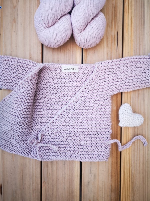 Zartrosa Wickeljäckchen in Krausrechts mit Wollknäueln und kleinem Strickherz auf Holz