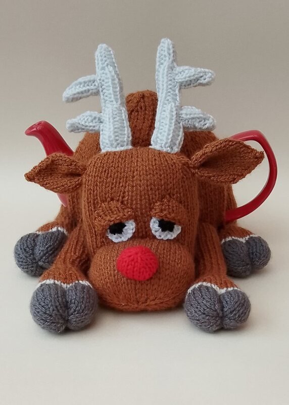 Brown knitted reindeer teapot cozy with a red nose, pale antlers, and grey hooves covering a red teapot.