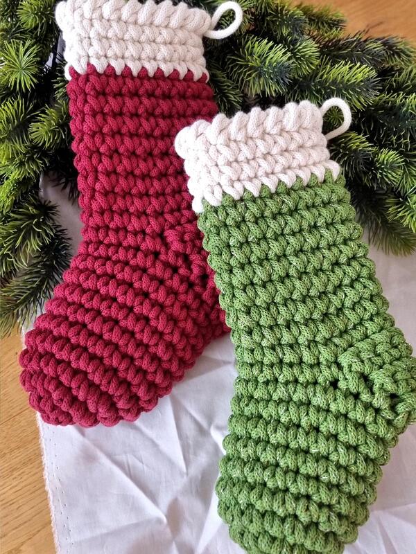 Zwei gehäkelte Weihnachtsstrümpfe in Rot und Grün mit cremefarbenen Bündchen und Aufhängeschlaufen vor Tannengrün.