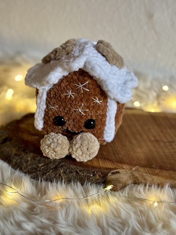 Gehäkeltes Lebkuchenhäuschen-Amigurumi mit weißem Zuckerguss, schwarzen Augen und Pompon-Füßen auf Holzscheibe vor Lichterkette