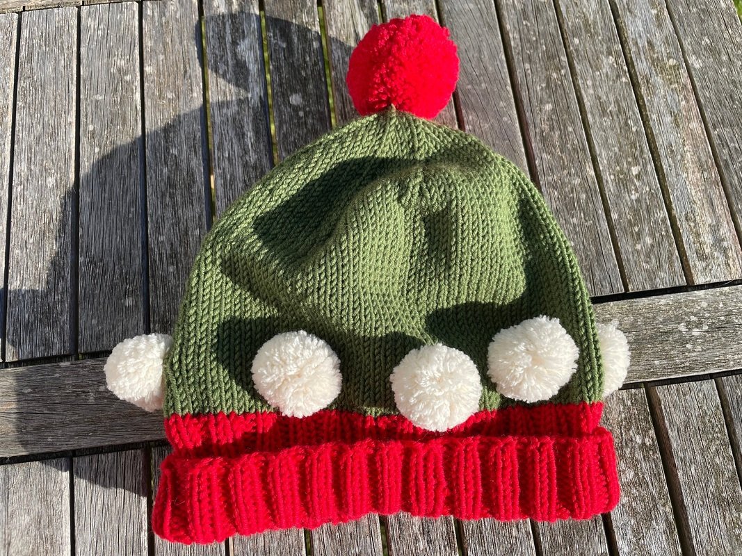 Gestrickte grün-rote Mütze mit rotem Bommel oben und weißen Bommeln am Saum auf Holzbrett