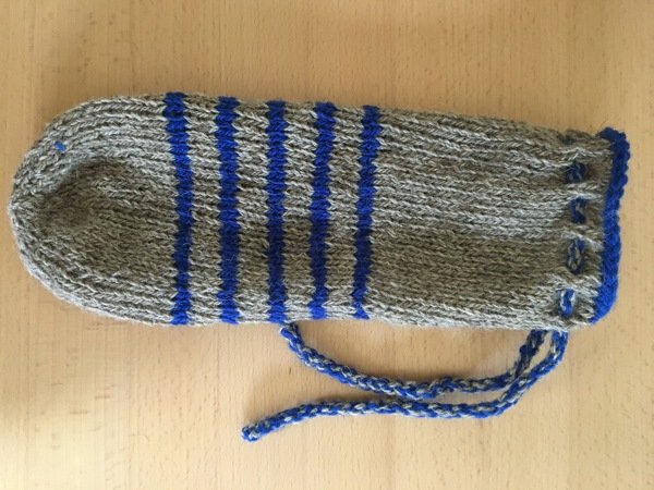 Gestrickte Wärmflaschenhülle in Grau mit blauen vertikalen Streifen und blauem Kordelzug auf Holzoberfläche