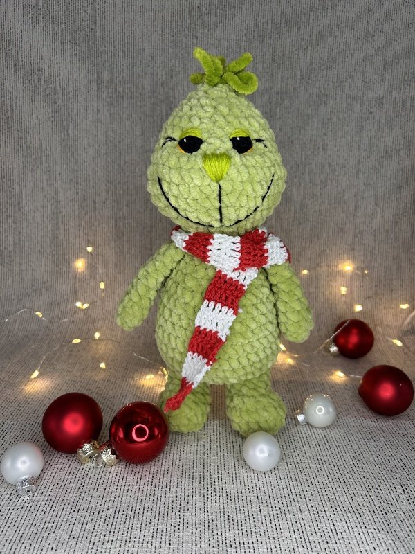Hellgrünes gehäkeltes Amigurumi mit rot-weißem Schal vor Lichterkette und Weihnachtskugeln