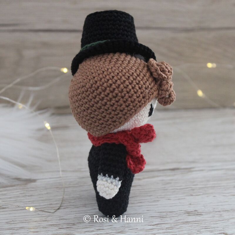 Gehäkelte Amigurumi-Figur mit schwarzem Zylinder und rotem Schal vor hellem Holz und Lichterkette