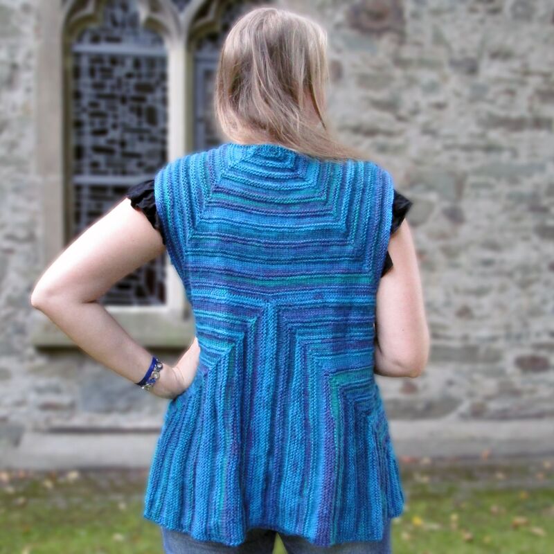 Back view of a woman wearing a blue, purple, and green variegated knitted sleeveless vest with a geometric back pattern.