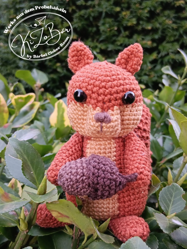 Gehäkeltes Amigurumi-Eichhörnchen (rostrot) sitzt in Blättern und hält eine gehäkelte Nuss
